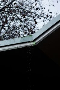 Rainwater flows from a shingled roof through a clean gutter and downspout beside a residential home.